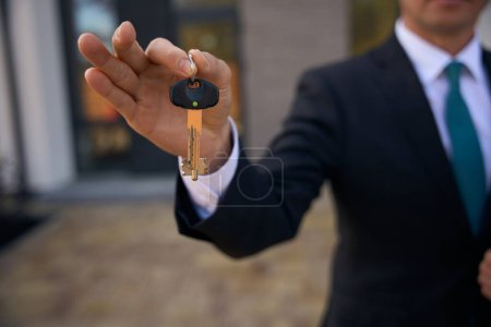 Foto de Foto de cerca del hombre de negocios en traje sosteniendo las llaves con dos dedos de la mano derecha mientras está de pie cerca de la construcción - Imagen libre de derechos