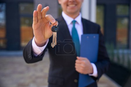 Téléchargez les photos : Photo recadrée de l'agent immobilier masculin montrant les clés dans une main et tenant presse-papiers dans un autre - en image libre de droit