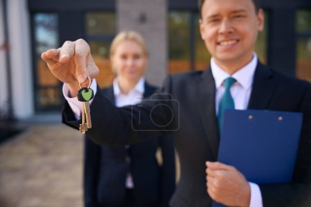 Téléchargez les photos : Photo recadrée d'un agent immobilier souriant tenant les clés avec deux doigts et une cliente debout derrière lui près du bâtiment - en image libre de droit
