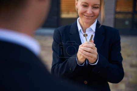 Cropped photo of amused woman in suit looking at keys in her hands and standing near the building