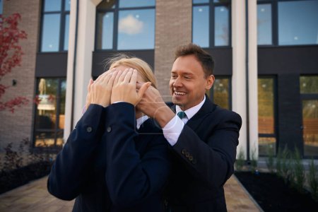 Téléchargez les photos : Homme joyeux debout derrière la femme et couvert ses yeux avec ses mains avant de montrer leur nouvelle maison - en image libre de droit
