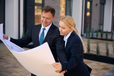 Photo for Waist up photo of man and lady standing while holding building plan in hands and looking at it - Royalty Free Image