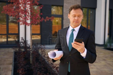Téléchargez les photos : Photo recadrée d'un agent immobilier debout près d'un immeuble avec du papier enroulé sous son bras et tapant sur un smartphone - en image libre de droit
