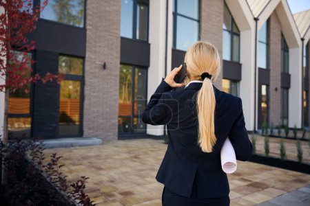 Téléchargez les photos : Vue de dos image de femme architecte debout devant les maisons tout en parlant au téléphone et tenant plan enroulé - en image libre de droit