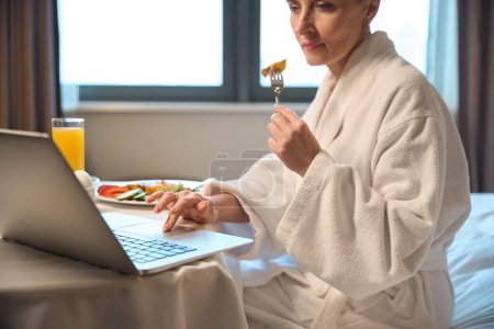 Photo for Cropped focused adult caucasian woman having breakfast and using laptop on bed at table in hotel room at morning. Concept of rest, vacation and travelling - Royalty Free Image