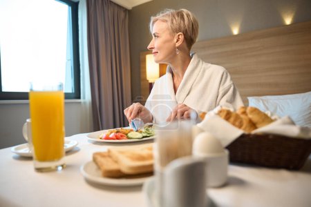 Foto de Mujer caucásica adulta sonriente con albornoz mirando hacia otro lado y desayunando en la cama a la mesa en la habitación del hotel por la mañana. Concepto de descanso, vacaciones y viajes - Imagen libre de derechos