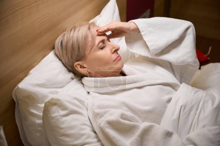 Exhausted adult caucasian woman wearing bathrobe lying and touching her head on bed in hotel room with lighting. Concept of rest, vacation and travelling