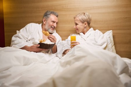 Photo for Smiling adult caucasian couple laying, resting and looking at each other on bed in hotel room with lighting. Concept of rest, vacation and travelling. Idea of relationship - Royalty Free Image