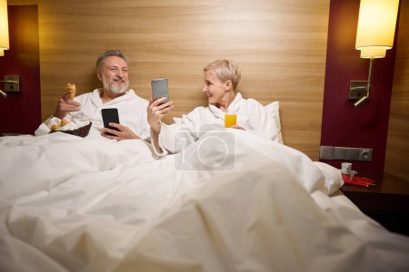 Foto de Sonriente pareja de mujeres caucásicas adultas mostrando algo en el teléfono inteligente a su marido en la cama en la habitación de hotel con iluminación. Concepto de descanso, vacaciones y viajes. Idea de relación - Imagen libre de derechos