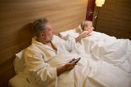 Foto de Hombre caucásico adulto con teléfono inteligente despertando a su esposa en la cama en la habitación de hotel con iluminación. Concepto de descanso, vacaciones y viajes. Idea de relación - Imagen libre de derechos