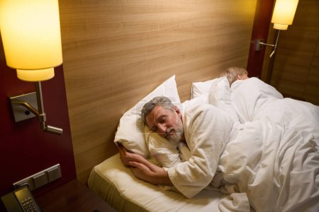 Téléchargez les photos : Couple européen d'âge moyen portant des peignoirs dormant sur le lit dans une chambre d'hôtel avec éclairage. Concept de repos, vacances et voyages. Idée de relation - en image libre de droit