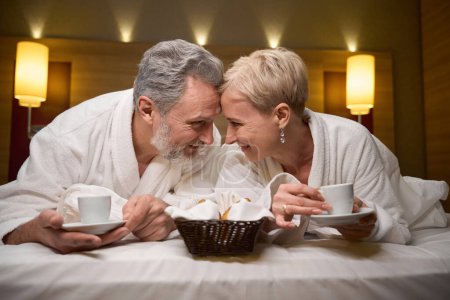 Téléchargez les photos : Vue de face de heureux couple caucasien adulte buvant du thé ou du café à partir de tasses et se regardant sur le lit dans la chambre d'hôtel avec éclairage. Concept de repos, vacances et voyages. Idée de relation - en image libre de droit