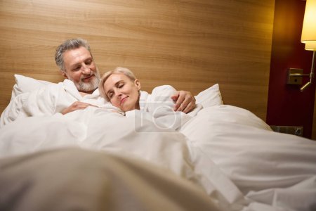 Foto de Hombre caucásico adulto abrazando a su esposa dormida en la cama en la habitación de hotel con iluminación. Concepto de descanso, vacaciones y viajes. Idea de relación - Imagen libre de derechos