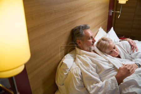 Téléchargez les photos : Vue en angle élevé du couple caucasien adulte portant des peignoirs embrassant et dormant sur le lit dans la chambre d'hôtel avec éclairage. Concept de repos, vacances et voyages. Idée de relation - en image libre de droit