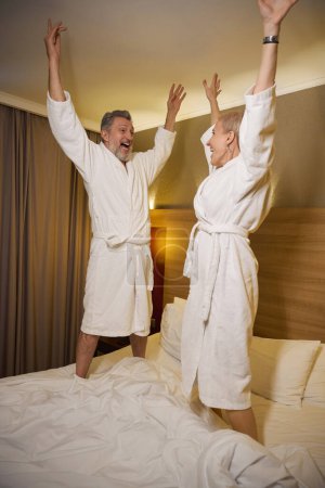 Téléchargez les photos : Joyeux couple caucasien adulte avec les mains dans l'air célébrant et se regardant tout en se tenant debout sur le lit dans la chambre d'hôtel avec éclairage. Concept de repos, vacances et voyages. Idée de relation - en image libre de droit