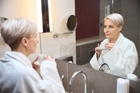 Téléchargez les photos : Femme caucasienne adulte portant un peignoir avec une tasse de thé ou de café se regardant dans un miroir dans la salle de bain le matin. Concept de style de vie féminin moderne - en image libre de droit