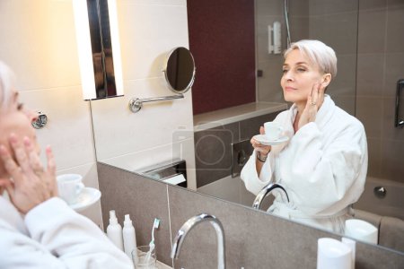 Téléchargez les photos : Femme caucasienne mature portant un peignoir avec une tasse de thé ou de café en regardant son visage dans un miroir dans la salle de bain le matin. Concept de style de vie féminin moderne - en image libre de droit