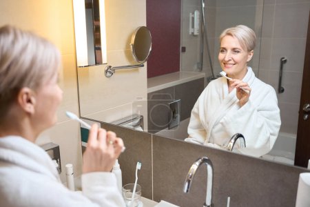 Foto de Mujer caucásica adulta sonriente usando albornoz cepillándose los dientes con cepillo de dientes mirándose en el espejo en el baño por la mañana. Concepto de procedimientos matutinos e higiene - Imagen libre de derechos