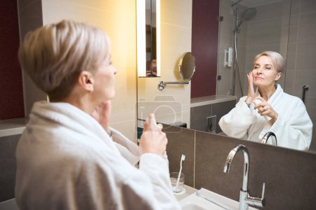 Téléchargez les photos : Femme caucasienne adulte concentrée portant un peignoir enduisant le visage de crème cosmétique et se regardant dans un miroir dans la salle de bain le matin. Concept de procédures matinales et d'hygiène - en image libre de droit