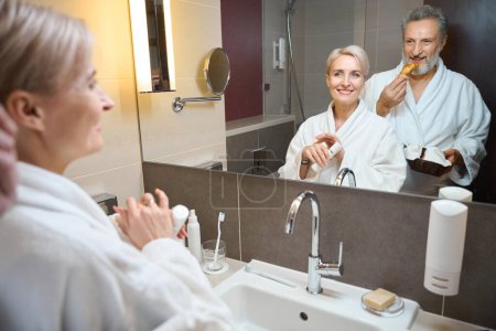 Photo for Smiling adult caucasian woman with cosmetic cream and man with croissants looking at themselves in mirror in bathroom at morning time. Concept of morning procedures and hygiene - Royalty Free Image