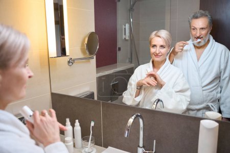 Téléchargez les photos : Femme caucasienne adulte avec crème cosmétique et homme brossant les dents avec une brosse à dents et ils se regardent dans le miroir dans la salle de bain le matin. Concept de procédures matinales et d'hygiène - en image libre de droit
