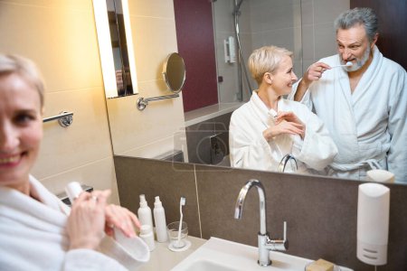 Téléchargez les photos : Miroir reflet de sourire couple caucasien adulte de la femme avec de la crème cosmétique en regardant l'homme brossant les dents avec une brosse à dents dans la salle de bain à l'heure du matin. Concept de procédures matinales et d'hygiène - en image libre de droit