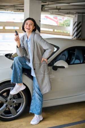 Téléchargez les photos : Joyeux jeune femme assise sur une nouvelle voiture, souriant et tenant les clés. Concept d'achat de transport - en image libre de droit