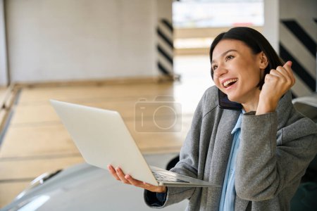 Foto de Señora de pie cerca de nuevo automóvil, sosteniendo el ordenador portátil y trabajando en línea, hablando por teléfono y se alegra. Concepto de negocio - Imagen libre de derechos