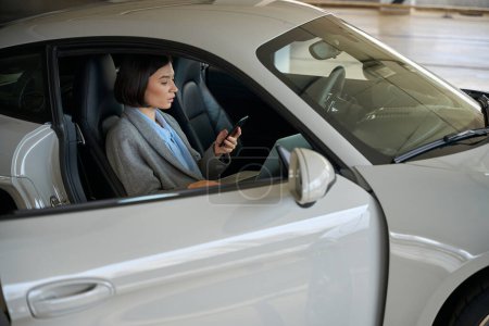 Photo for Female sitting in new car, holding smartphone and looking on screen, working online - Royalty Free Image
