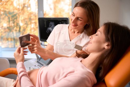 Téléchargez les photos : Femme enceinte gaie et médecin regardant la photo échographie à l'hôpital - en image libre de droit