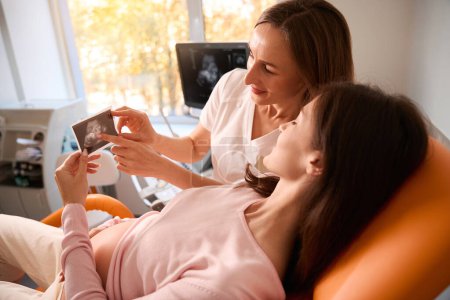 Téléchargez les photos : Jeune femme enceinte visitant le médecin à la clinique regardant la photo d'échographie à l'hôpital - en image libre de droit