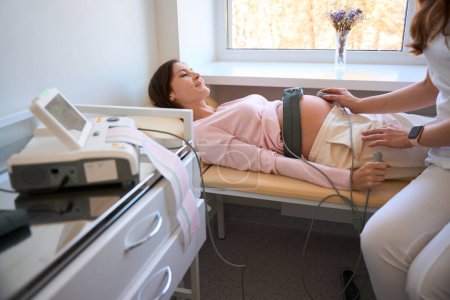 Téléchargez les photos : Femme enceinte pendant la cardiotocographie du fœtus pendant la grossesse et l'accouchement dans le centre prénatal - en image libre de droit