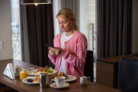 Téléchargez les photos : Focused dame regardant smartphone dans ses mains tout en étant assis à la table avec de la nourriture dans la chambre d'hôtel - en image libre de droit
