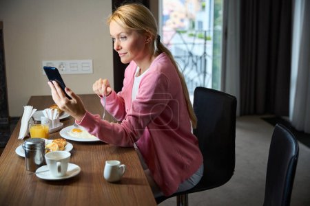 Téléchargez les photos : Focused dame regardant le téléphone cellulaire dans sa main tout en mangeant des œufs à table dans la suite - en image libre de droit