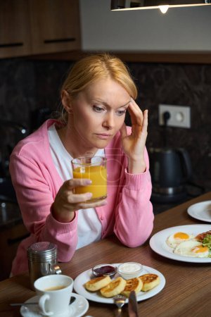 Téléchargez les photos : Triste femme coûteuse avec verre de boisson aux agrumes assis à la table de cuisine chargée de assiettes avec des aliments pour le petit déjeuner - en image libre de droit