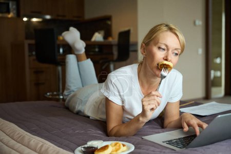 Foto de Señora acostada en la cama en el dormitorio comiendo panqueque pinchó en tenedor mientras miraba el monitor de la computadora - Imagen libre de derechos