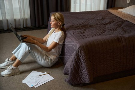 Photo for Focused woman typing on portable computer while sitting on carpeted floor near bed in hotel room - Royalty Free Image