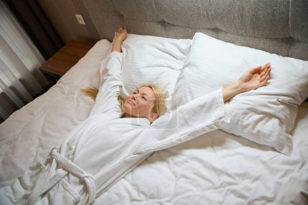 Téléchargez les photos : Dame satisfaite en peignoir couché couché couché sur le lit dans la chambre et faisant étirement du bras - en image libre de droit