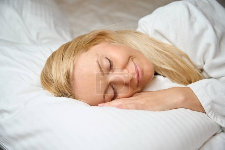 Photo for Serene woman dressed in bathrobe lying with eyes closed on soft white pillow in bed - Royalty Free Image
