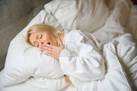 Photo for Sleepy woman in terry bathrobe lying on pillow in bed and yawning - Royalty Free Image