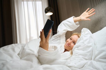 Téléchargez les photos : Dame satisfaite en peignoir agitant quelqu'un sur son écran de smartphone alors qu'elle était couchée au lit - en image libre de droit