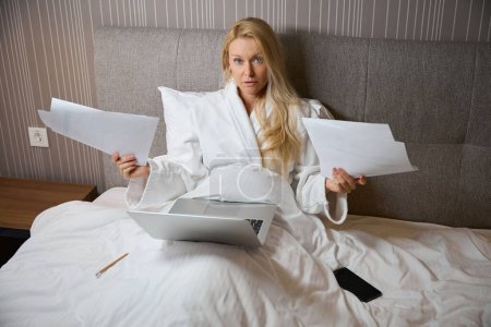Photo for Serious lady seated in bed in suite in front of laptop holding pile of documents in hands - Royalty Free Image