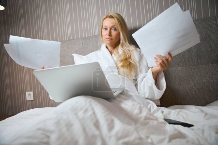 Photo for Serious business lady sitting in bed in hotel room with portable computer and pile of documents in her hands - Royalty Free Image