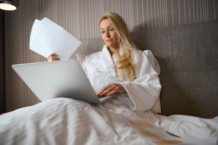 Photo for Focused businesswoman seated in bed in suite with pile of documents in her hand typing on laptop - Royalty Free Image