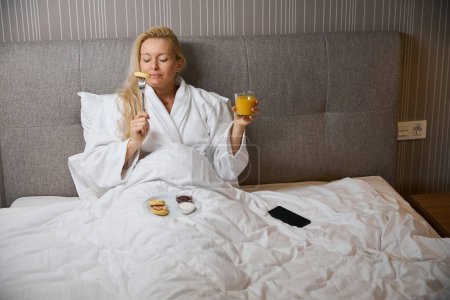 Téléchargez les photos : Dozy femelle assise dans son lit tenant un verre de jus d'orange à la main soulevant la crêpe piquée sur la fourchette à sa bouche - en image libre de droit