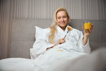 Photo for Contented lady seated in bed in suite holding glass of orange juice and pancake pricked on fork in hands - Royalty Free Image