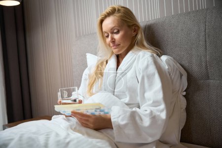 Foto de Mujer enfocada sentada en la cama en suite sosteniendo un vaso de agua y mirando el pillbox en sus manos - Imagen libre de derechos