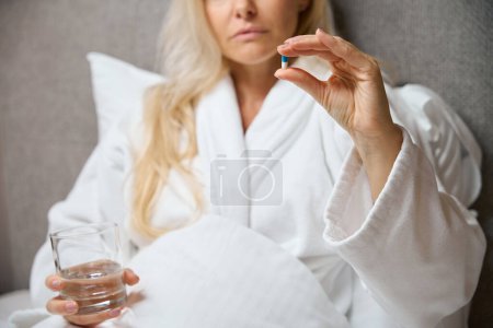 Cropped photo of female in bathrobe seated in bed holding glass of water and drug capsule in hands