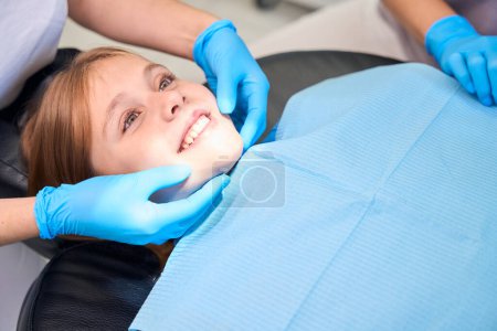 Téléchargez les photos : Adolescente souriante placée sur une chaise pour les procédures dentaires, personnel médical travaillant dans des gants de protection - en image libre de droit