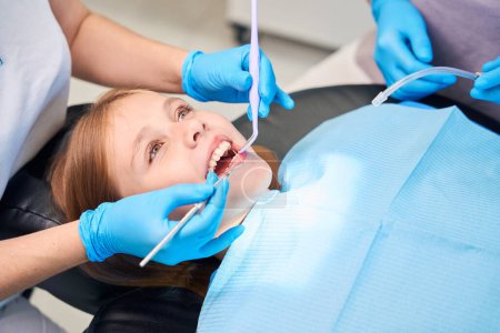 Téléchargez les photos : Jeune patiente dans la chaise des dentistes, le personnel médical utilise des outils spéciaux au travail - en image libre de droit